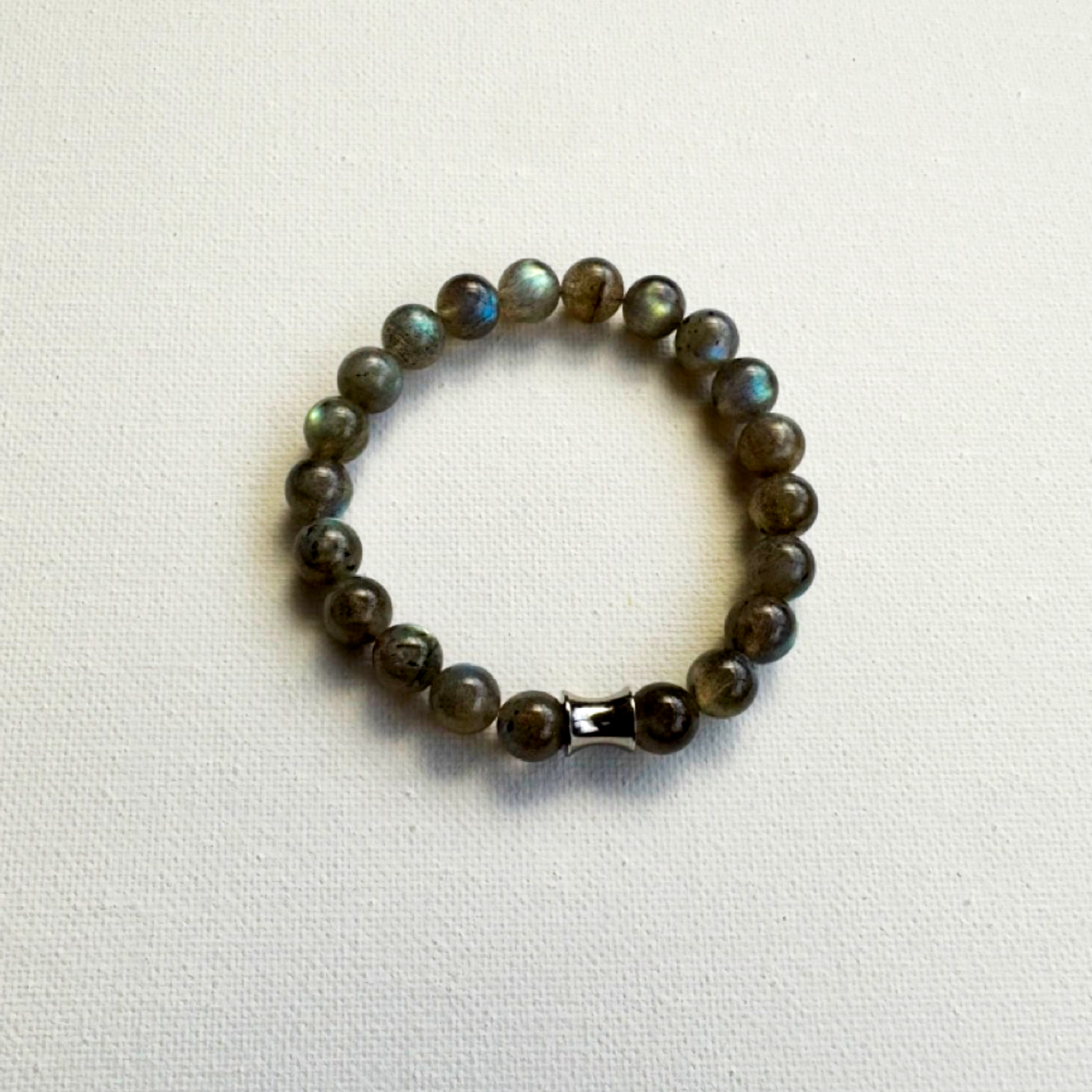 A beaded stretch bracelet made of labradorite beads with a shimmering green-blue iridescent finish, featuring a polished silver-toned metal focal, displayed on a textured off-white fabric background.