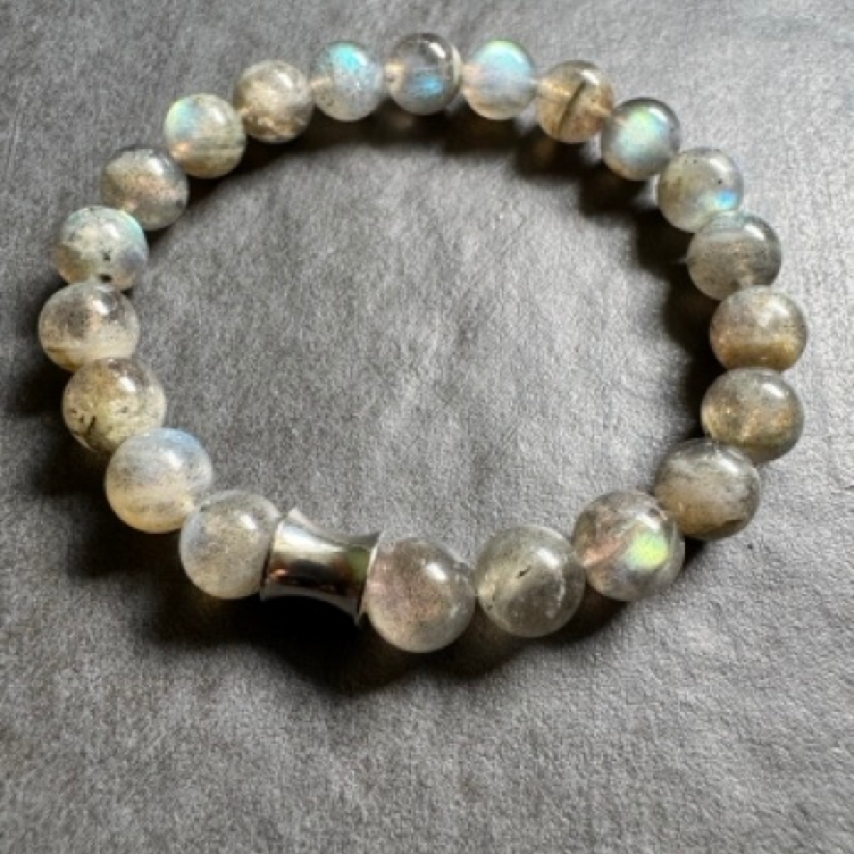 A beaded stretch bracelet made of round, polished labradorite beads with an iridescent silver sheen, featuring a small metallic silver-toned accent bead, displayed on a textured gray surface.