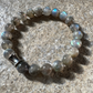 A beaded stretch bracelet made of round gray labradorite beads with an iridescent sheen and a silver-toned cylindrical accent bead, photographed on a textured stone surface.