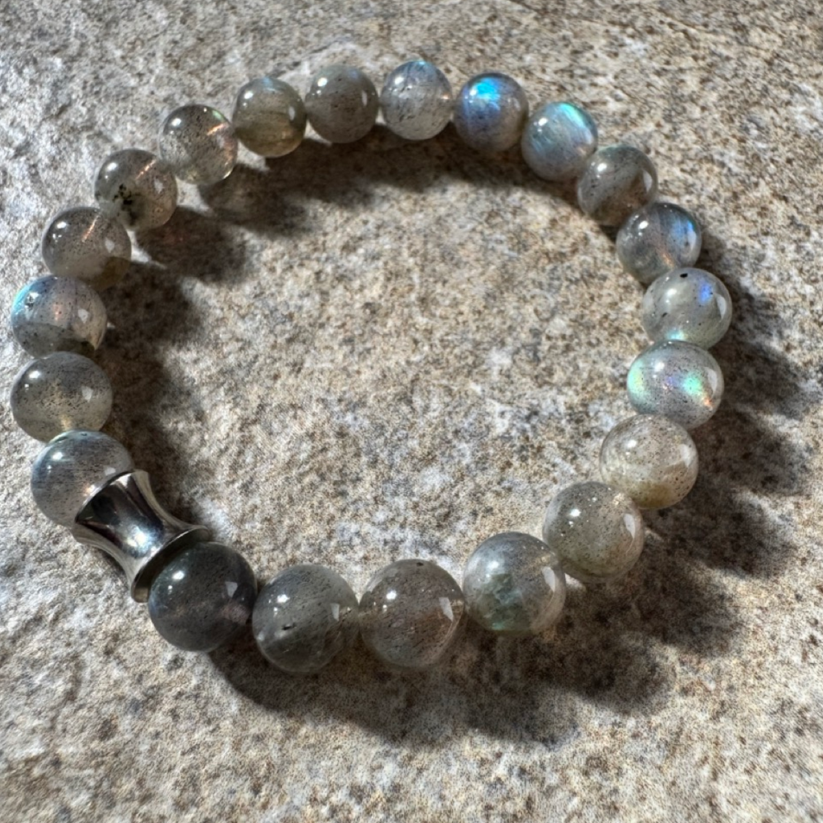 A beaded stretch bracelet made of round gray labradorite beads with an iridescent sheen and a silver-toned cylindrical accent bead, photographed on a textured stone surface.