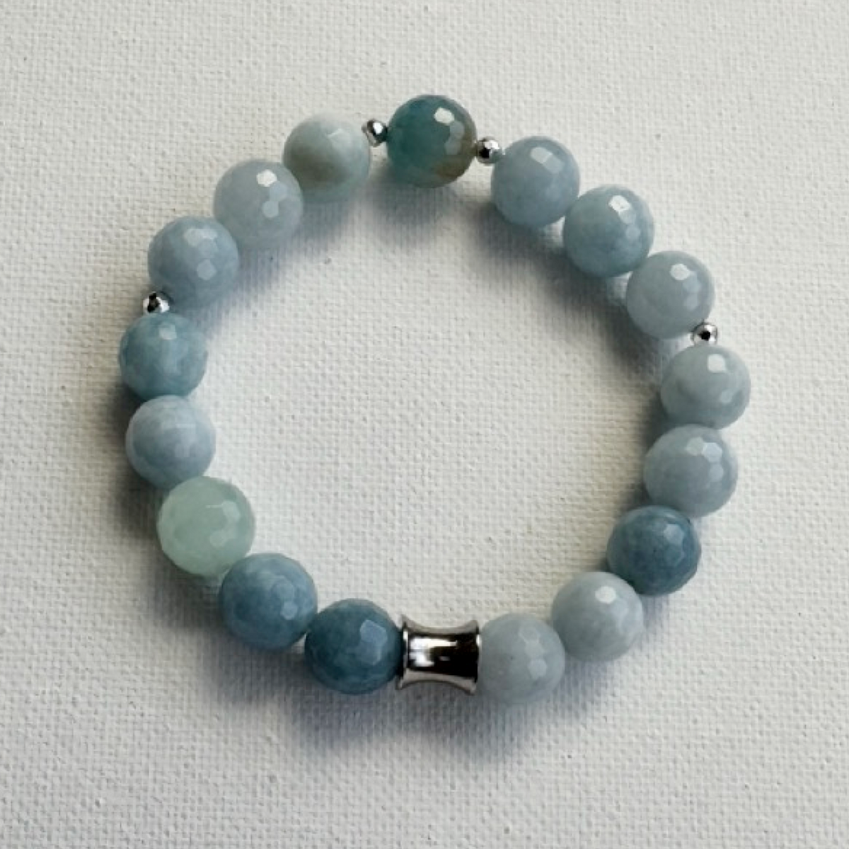 A beaded stretch bracelet made of faceted light blue and pale green gemstone beads, accented by small silver spacer beads and a silver cylindrical clasp, displayed on a textured off-white fabric background.