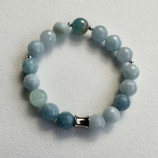 A beaded stretch bracelet made of faceted light blue and pale green gemstone beads, accented by small silver spacer beads and a silver cylindrical clasp, displayed on a textured off-white fabric background.
