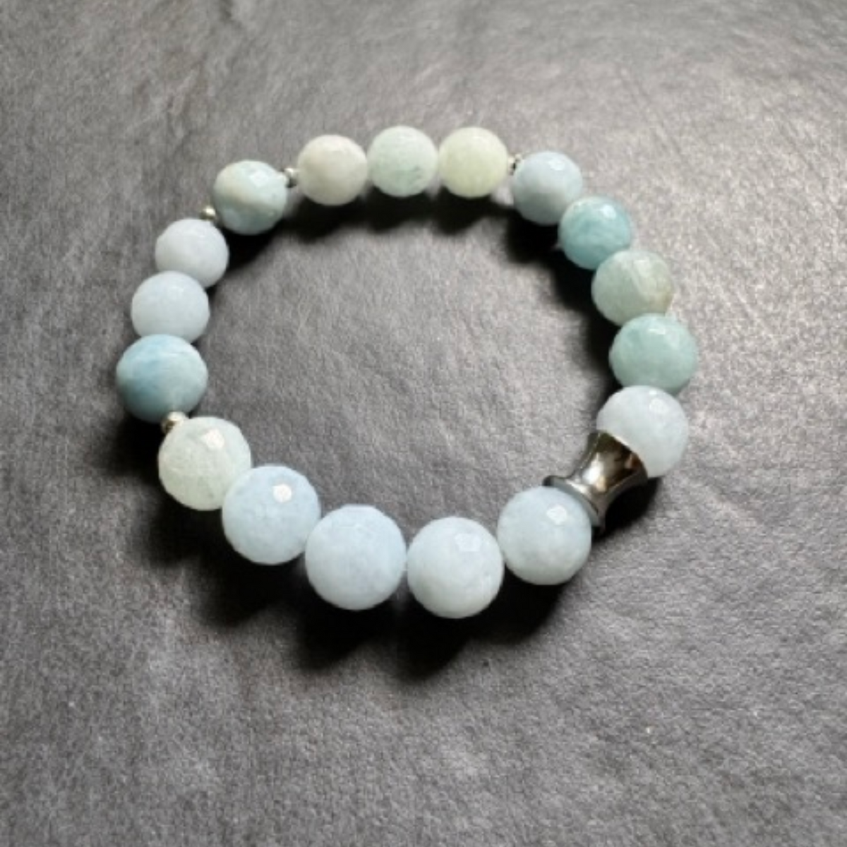 A beaded stretch bracelet made of light blue and white round beads, featuring a silver-toned metal accent bead, displayed on a dark gray textured surface.