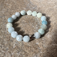 A beaded stretch bracelet made of faceted round beads in pale blue, white, and light green, with a polished silver-toned cylindrical connector, displayed on a textured gray stone surface.