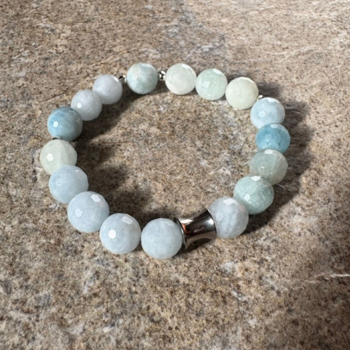 A beaded stretch bracelet made of faceted round beads in pale blue, white, and light green, with a polished silver-toned cylindrical connector, displayed on a textured gray stone surface.