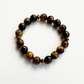 A beaded stretch bracelet made of polished tiger's eye beads in brown and black, accented by small metallic gold spacer beads, displayed on a clean white textured background.