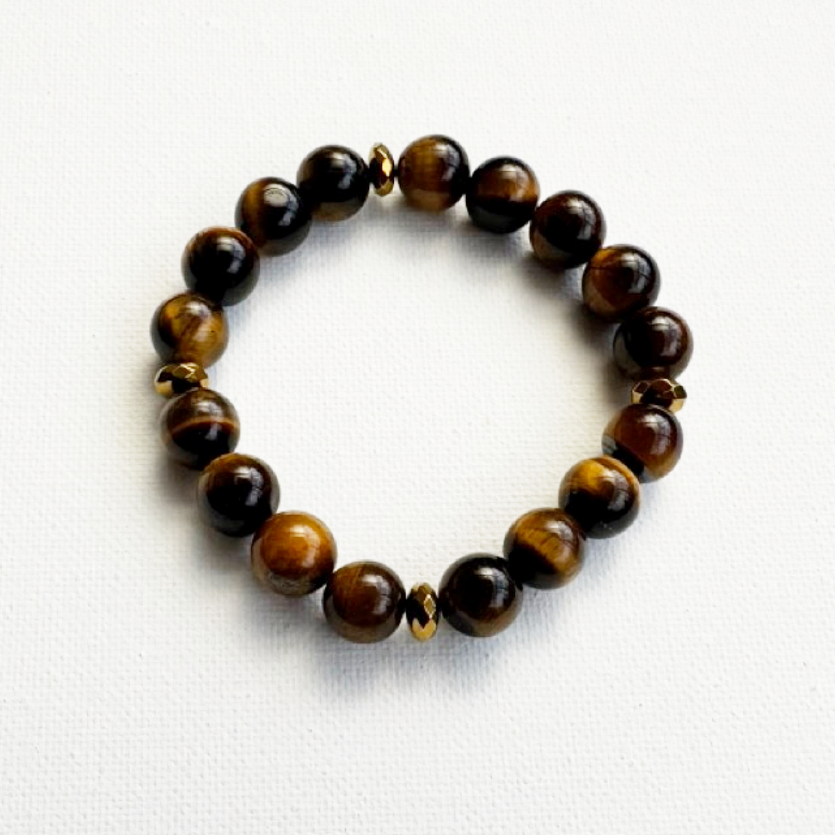 A beaded stretch bracelet made of polished tiger's eye beads in brown and black, accented by small metallic gold spacer beads, displayed on a clean white textured background.