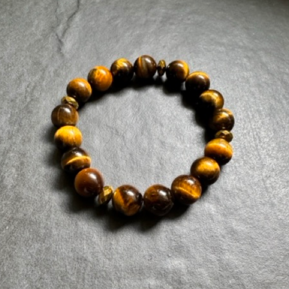 A beaded stretch bracelet made of polished tiger's eye stones, arranged in a circular shape on a textured gray slate surface.