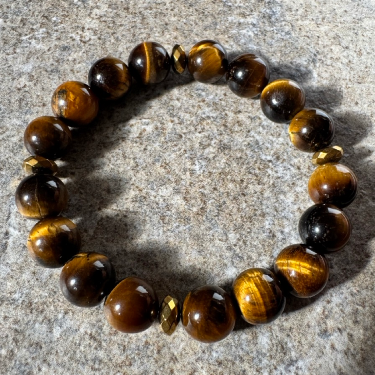 A beaded stretch bracelet made of polished tiger's eye beads with small faceted gold spacer beads, displayed on a textured gray stone surface.