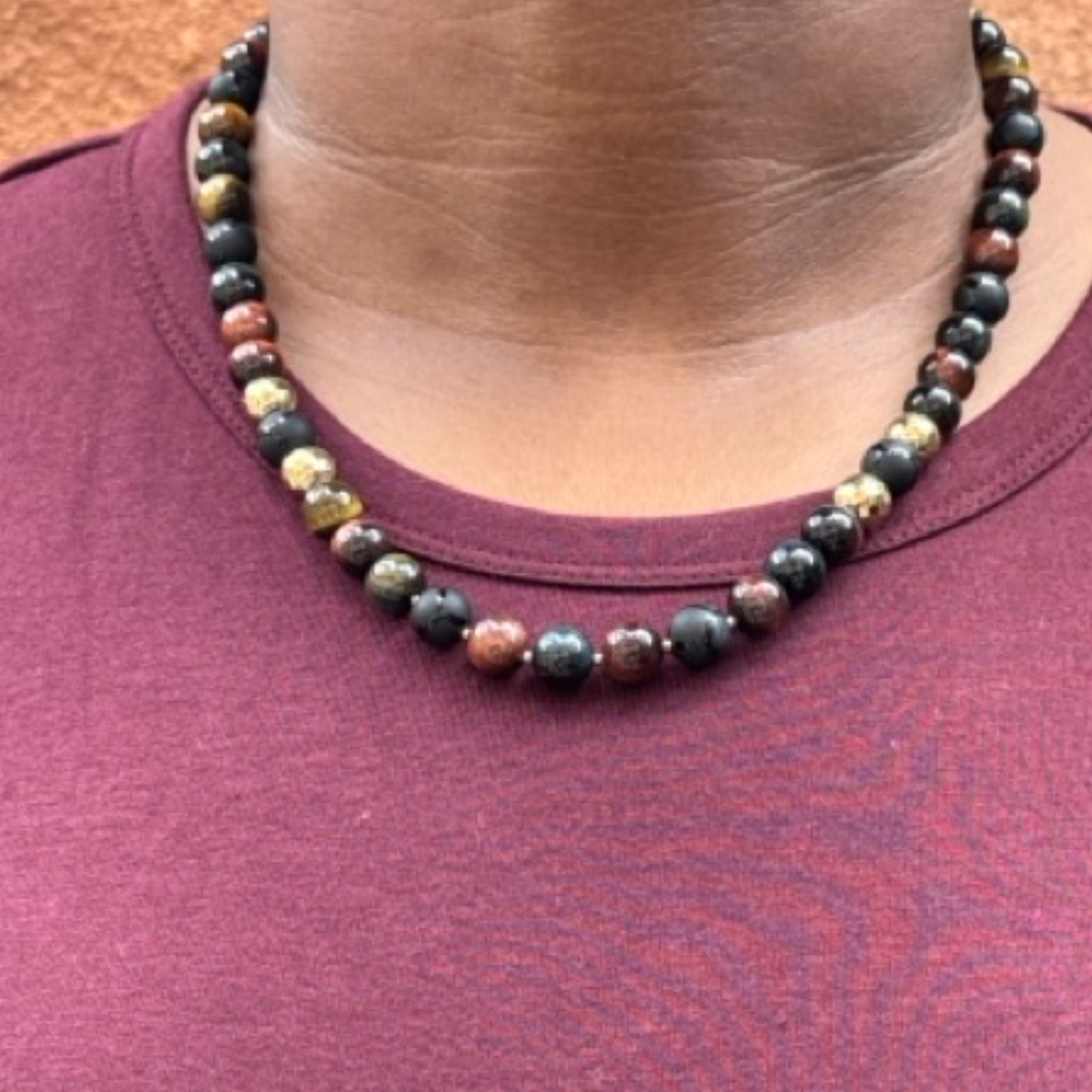 A close-up of a person wearing a multi-colored beaded necklace with dark, metallic, and gold-toned beads, paired with a maroon t-shirt.