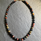 A beaded necklace made of polished round gemstone beads in black, brown, orange, and gold, arranged in an oval on a textured gray slate background.