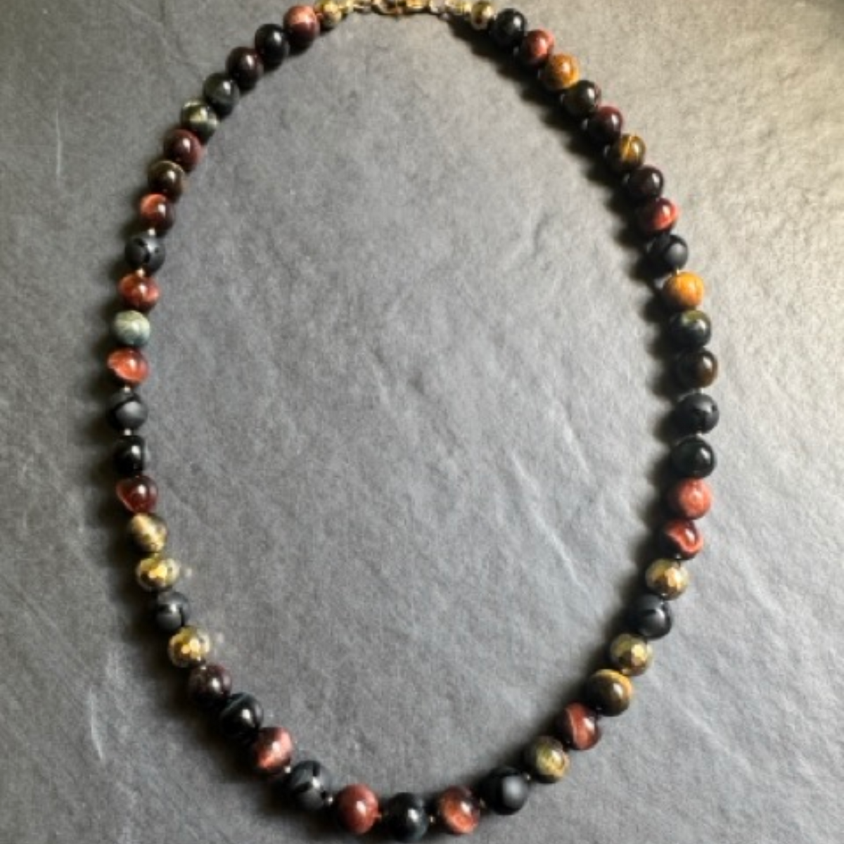A beaded necklace made of polished round gemstone beads in black, brown, orange, and gold, arranged in an oval on a textured gray slate background.