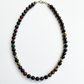 A beaded necklace made of polished round dark brown and black gemstone beads with small metallic gold spacer beads and a gold clasp, displayed on a clean white background.