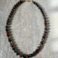 A beaded necklace made of polished round gemstone beads in black, red, brown, and metallic gold, arranged in an oval on a textured light beige stone surface.