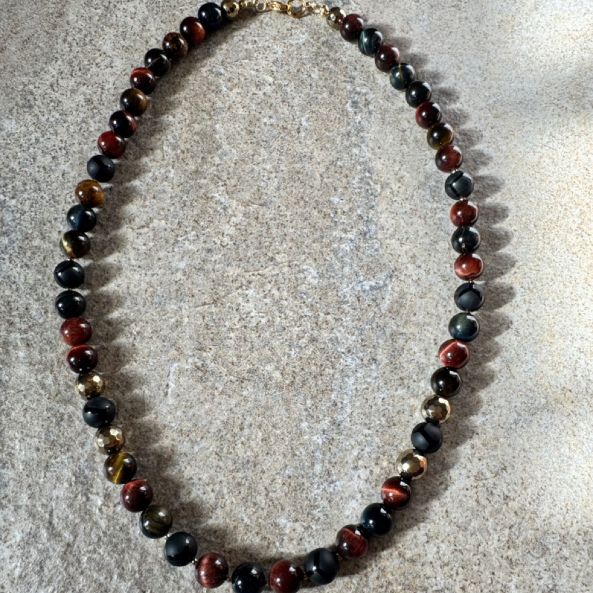 A beaded necklace made of polished round gemstone beads in black, red, brown, and metallic gold, arranged in an oval on a textured light beige stone surface.