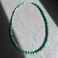 A beaded necklace made of small turquoise-green and blue marbled beads with a silver clasp, displayed on a gray textured surface with dramatic shadows.