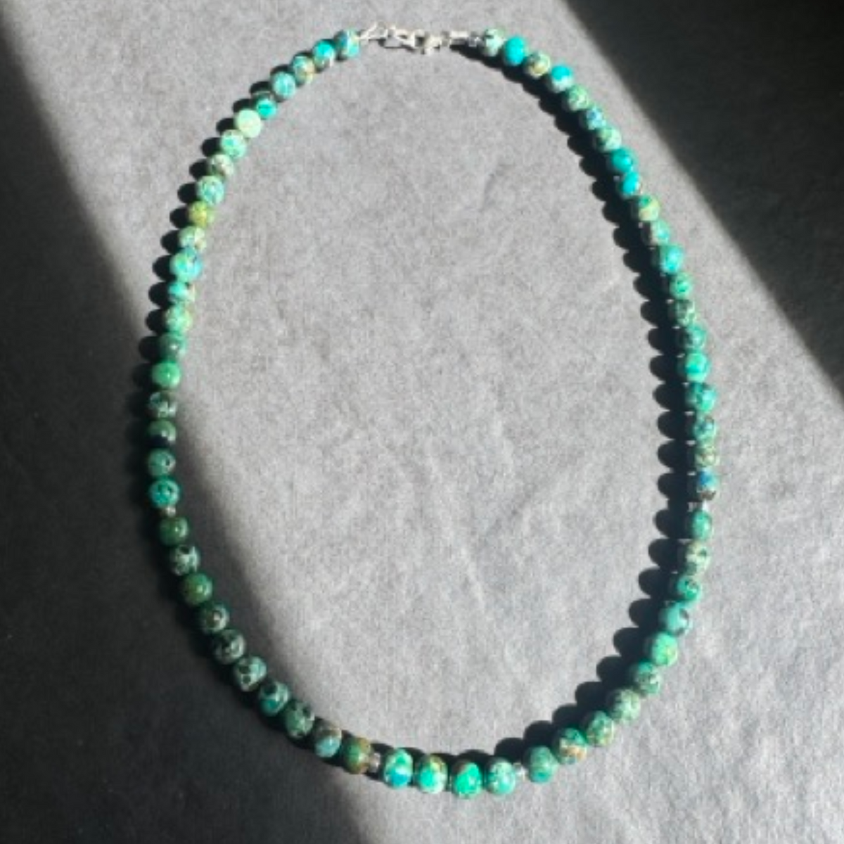 A beaded necklace made of small turquoise-green and blue marbled beads with a silver clasp, displayed on a gray textured surface with dramatic shadows.