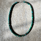 A beaded necklace made of small turquoise-green and dark brown marbled beads, arranged in an oval shape on a textured gray stone surface.