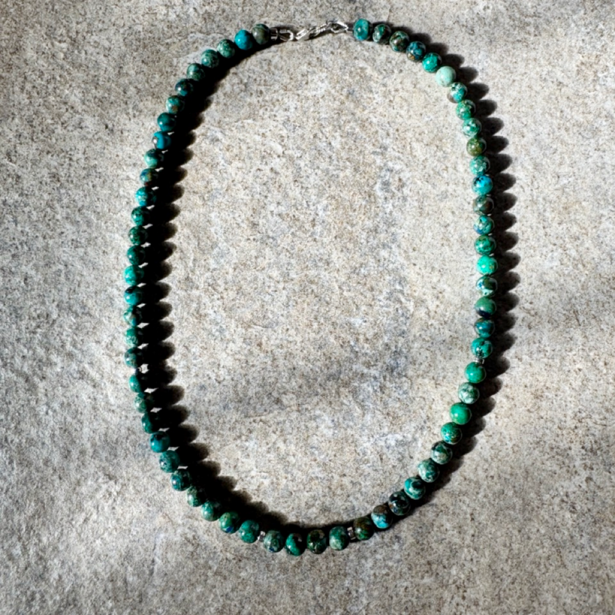 A beaded necklace made of small turquoise-green and dark brown marbled beads, arranged in an oval shape on a textured gray stone surface.