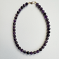A beaded necklace made of faceted dark purple beads separated by small gold spacer beads, with a gold clasp, laid flat on a light gray background.