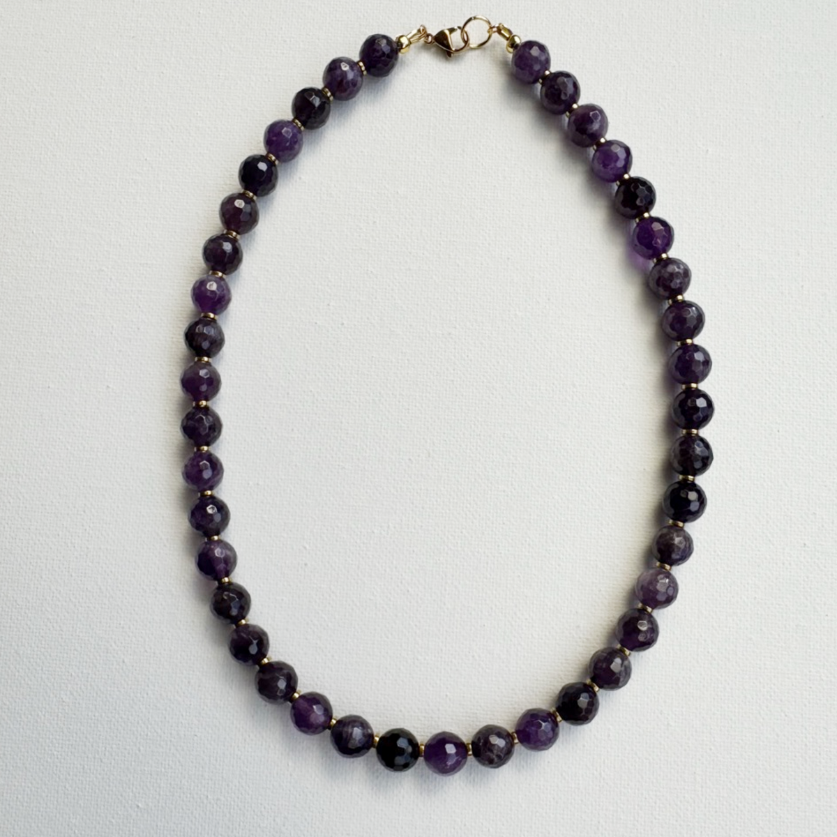 A beaded necklace made of faceted dark purple beads separated by small gold spacer beads, with a gold clasp, laid flat on a light gray background.