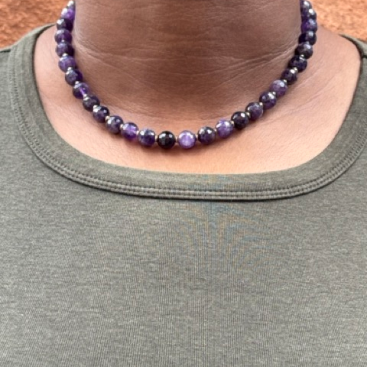 A close-up of a person wearing a beaded choker necklace made of round purple and dark violet beads, paired with an olive green crew-neck shirt.