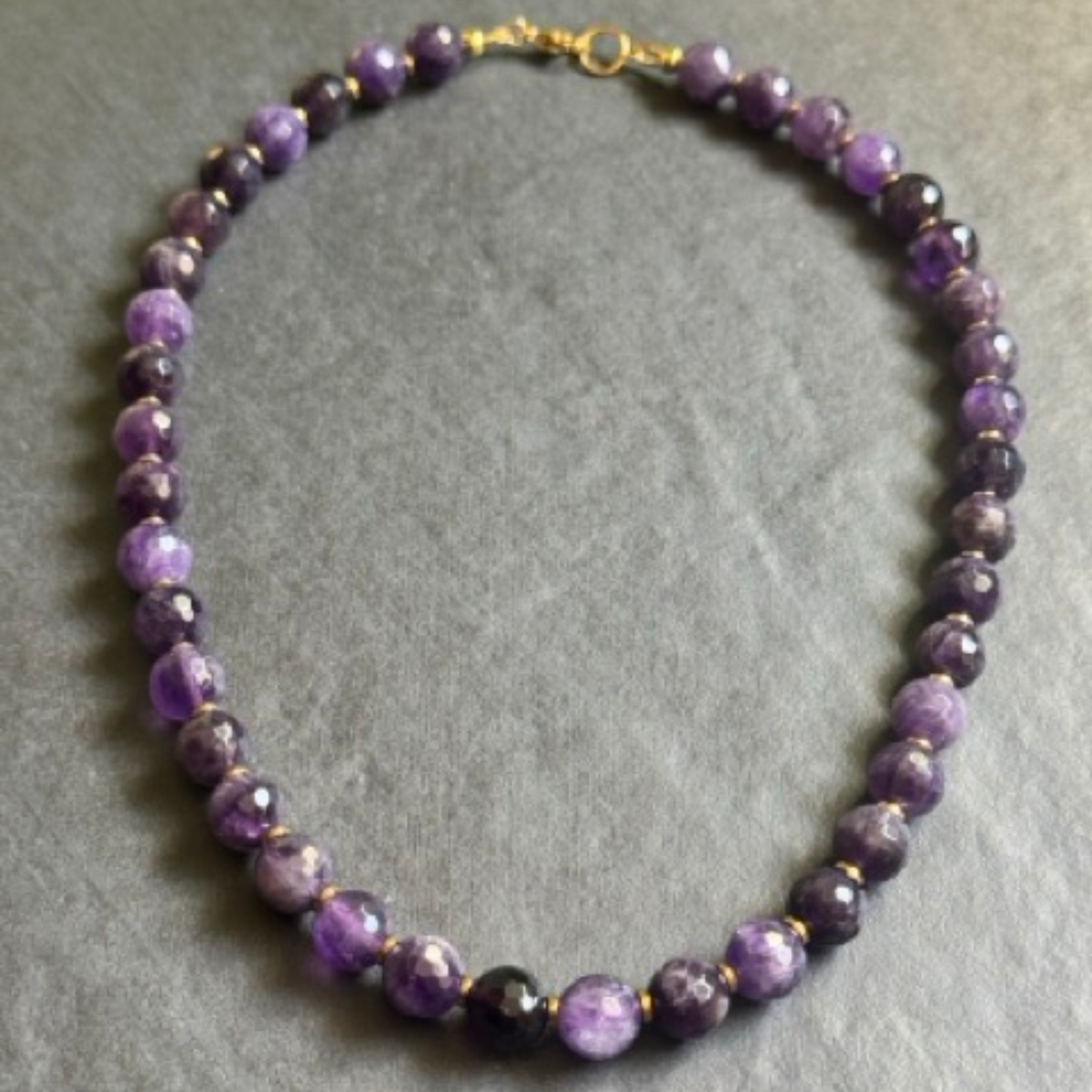 A beaded necklace made of faceted purple and violet beads with small gold spacer beads and a gold clasp, displayed on a textured gray background.