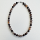A beaded necklace made of round agate-like beads in dark brown, black, gray, and light brown marbled patterns, arranged in an oval on a white textured background.