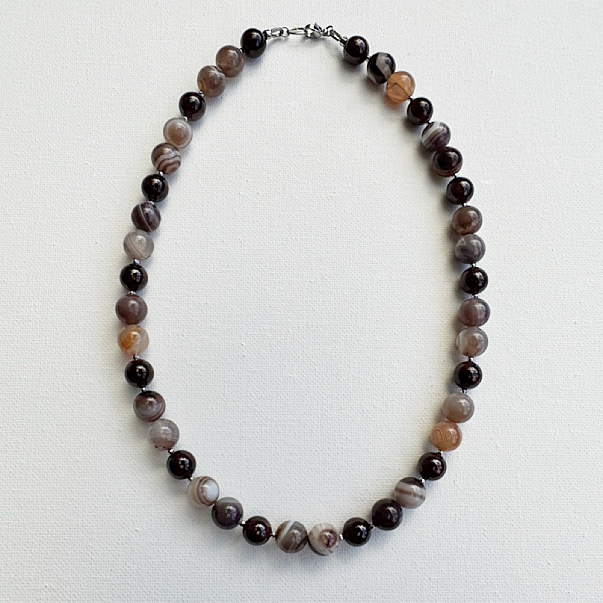 A beaded necklace made of round agate-like beads in dark brown, black, gray, and light brown marbled patterns, arranged in an oval on a white textured background.