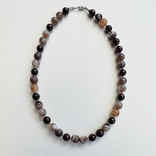 A beaded necklace made of round agate-like beads in dark brown, black, gray, and light brown marbled patterns, arranged in an oval on a white textured background.