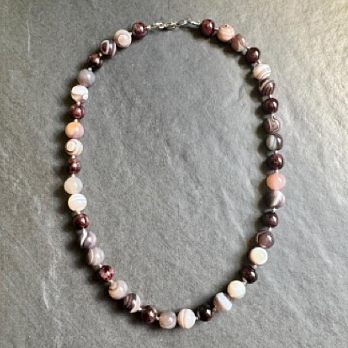 A beaded necklace made of round marbled beads in shades of burgundy, pink, white, and gray, arranged in a circle on a textured gray slate background.