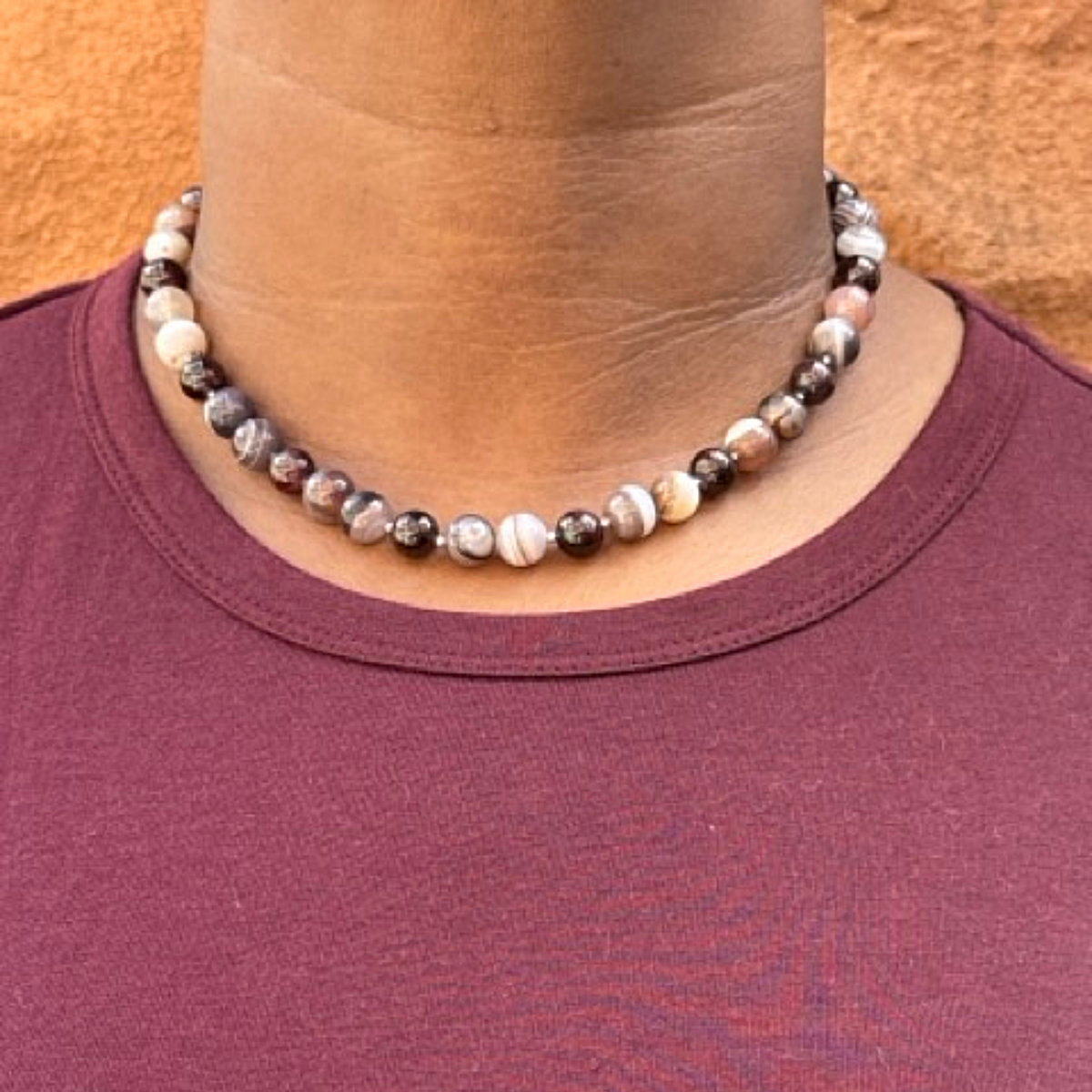 A close-up of a person wearing a beaded necklace made of marbled beads in white, gray, brown, and black, paired with a burgundy t-shirt against a textured orange wall.