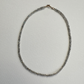 A delicate beaded necklace made of small gray faceted beads with a rose gold clasp, laid flat on a textured white background.