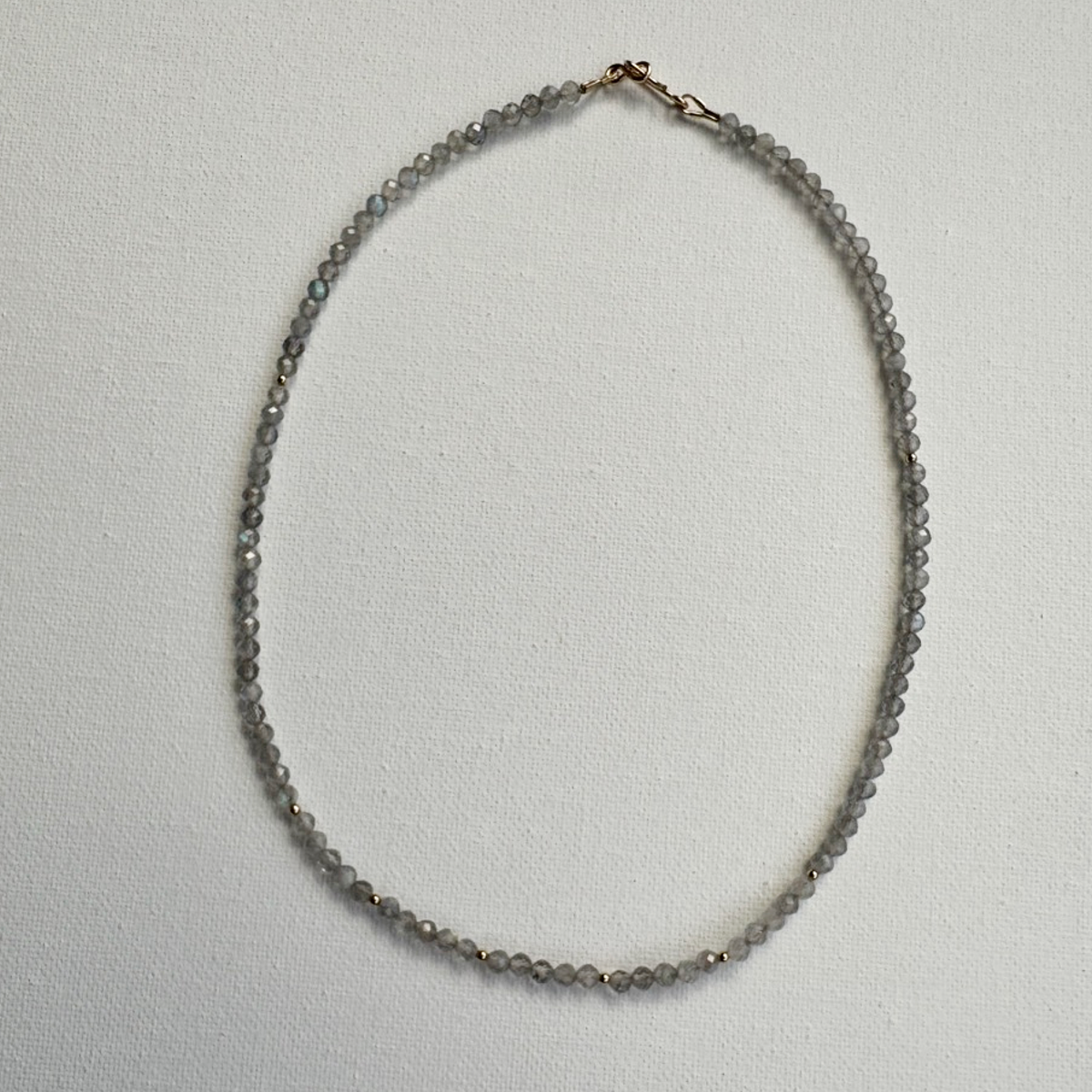 A delicate beaded necklace made of small gray faceted beads with a rose gold clasp, laid flat on a textured white background.