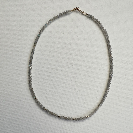 A delicate beaded necklace made of small gray faceted beads with a rose gold clasp, laid flat on a textured white background.