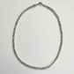 A delicate beaded necklace made of small gray faceted beads with a small gold clasp, displayed on a textured white background.