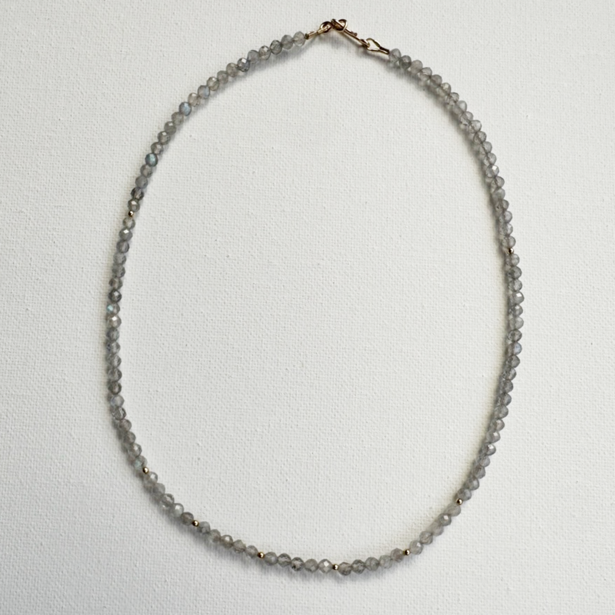 A delicate beaded necklace made of small gray faceted beads with a small gold clasp, displayed on a textured white background.