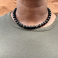A close-up of a person's neck and upper chest wearing a beaded necklace made of glossy black round beads, paired with an olive green crewneck shirt against a textured orange-brown background.