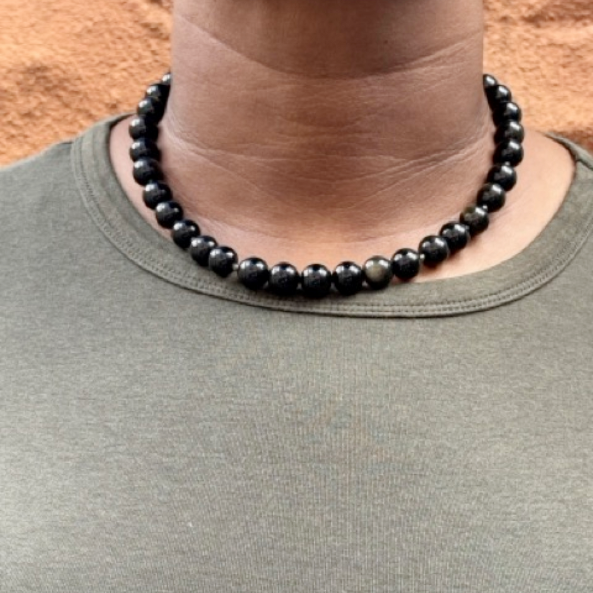 A close-up of a person's neck and upper chest wearing a beaded necklace made of glossy black round beads, paired with an olive green crewneck shirt against a textured orange-brown background.