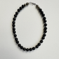 A beaded necklace made of glossy black round beads with a small section of dark gray textured beads and a silver-toned clasp, displayed on a light gray textured background.