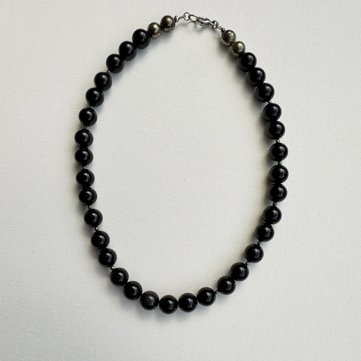 A beaded necklace made of glossy black round beads with a small section of dark gray textured beads and a silver-toned clasp, displayed on a light gray textured background.