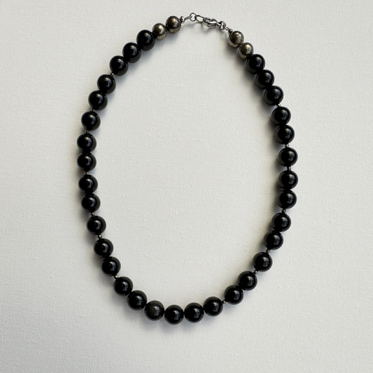 A beaded necklace made of glossy black round beads with a small section of dark gray textured beads and a silver-toned clasp, displayed on a light gray textured background.