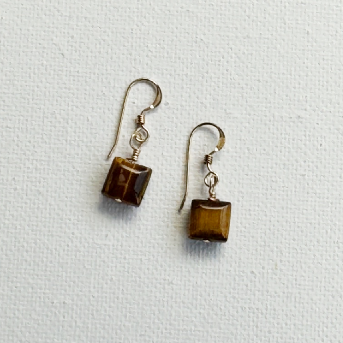 A pair of dangling earrings with square brown tiger's eye stones and gold-toned fishhook wires, displayed on a textured white background.