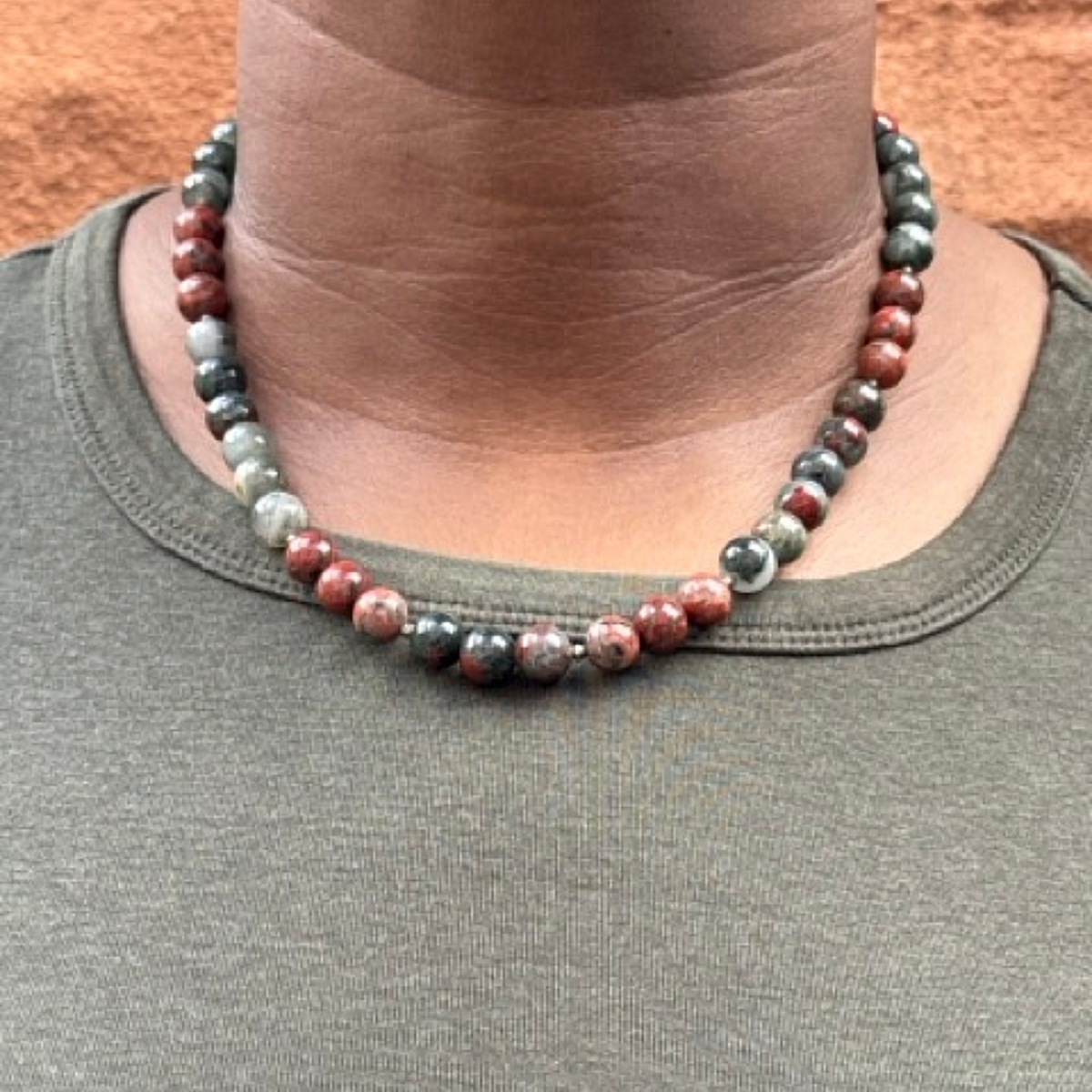 A close-up of a person wearing a beaded necklace made of polished round beads in red, gray, black, and white, paired with an olive green crewneck shirt.