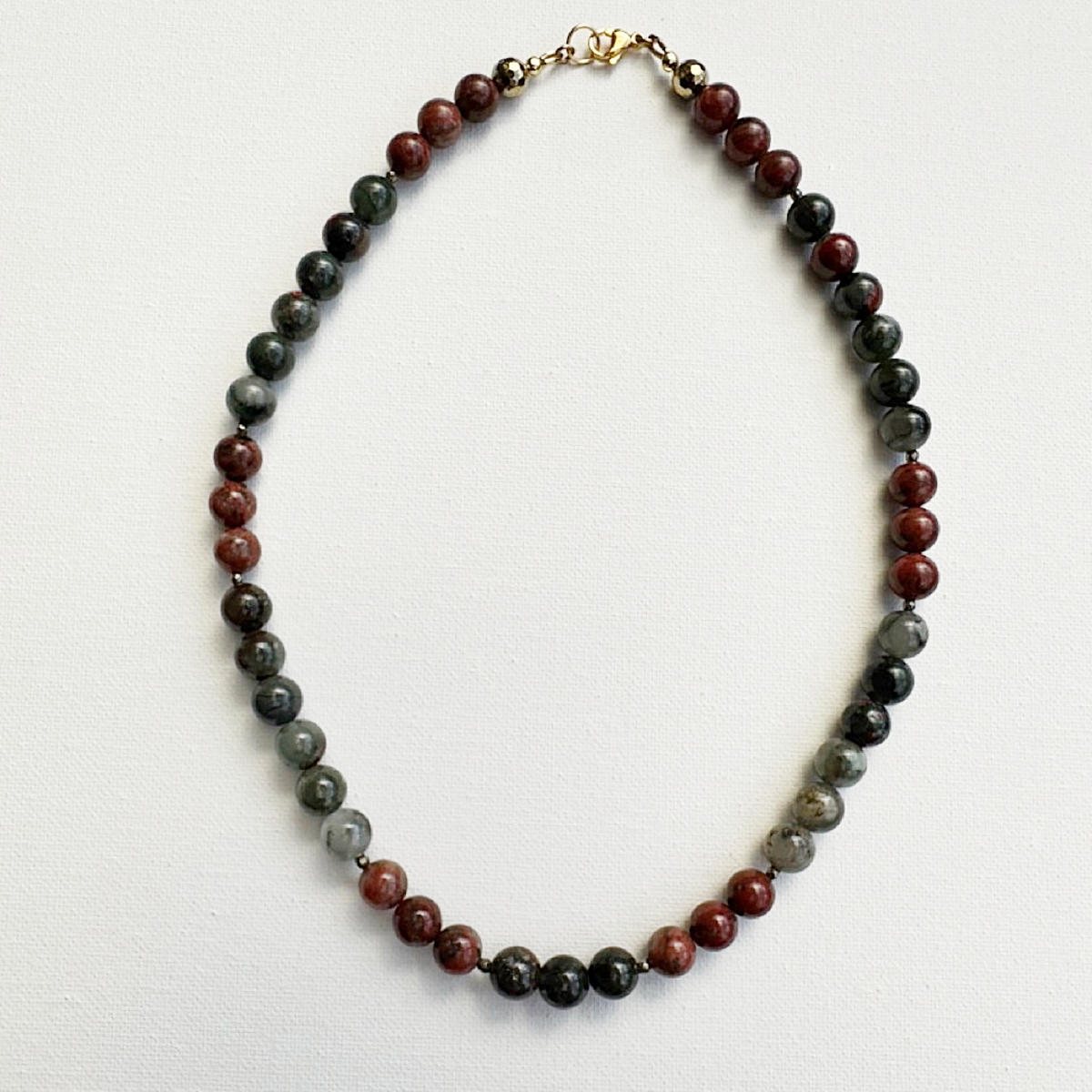 A beaded necklace made of alternating dark green, reddish-brown, and gray polished stone beads, finished with a gold clasp, laid flat on a white background.