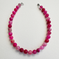 A beaded necklace made of faceted round pink gemstone beads in various shades of magenta and rose, arranged in a circle on a white textured background. The necklace has silver-toned metal clasps at the top.