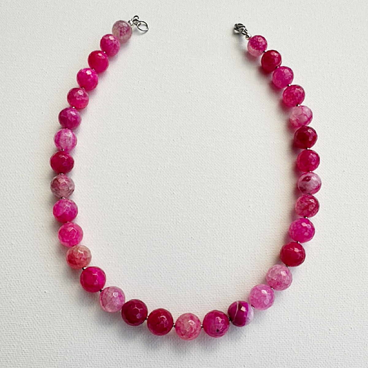 A beaded necklace made of faceted round pink gemstone beads in various shades of magenta and rose, arranged in a circle on a white textured background. The necklace has silver-toned metal clasps at the top.