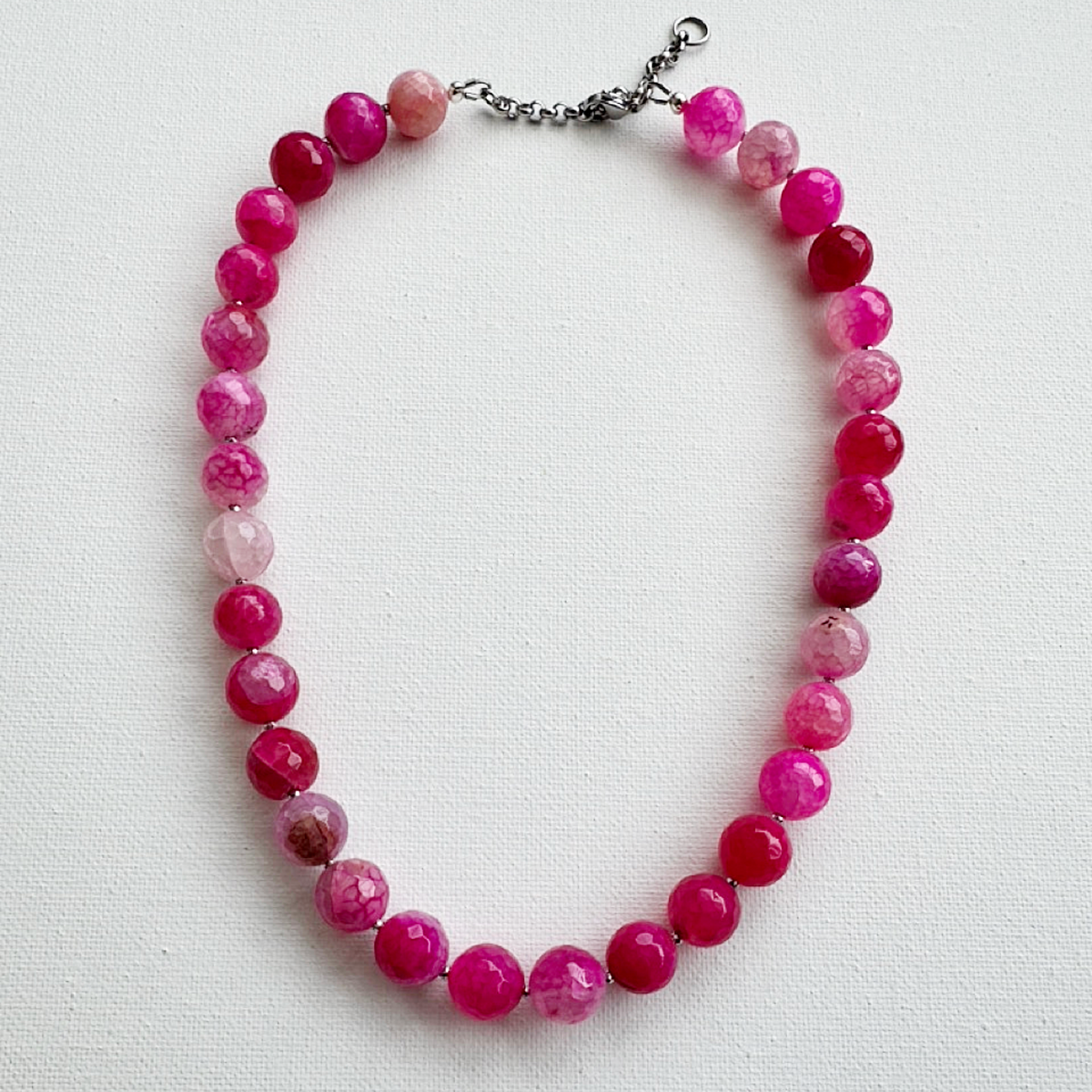 A beaded necklace made of faceted round beads in shades of pink, magenta, and translucent light pink, with a silver-tone chain clasp, laid flat on a white textured background.