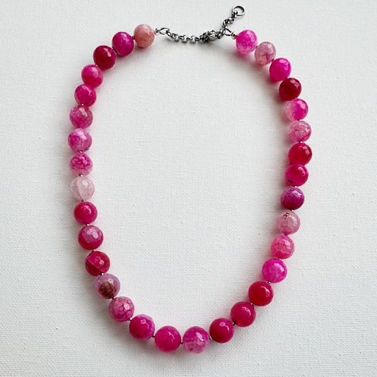 A beaded necklace made of faceted round beads in shades of pink, magenta, and translucent light pink, with a silver-tone chain clasp, laid flat on a white textured background.