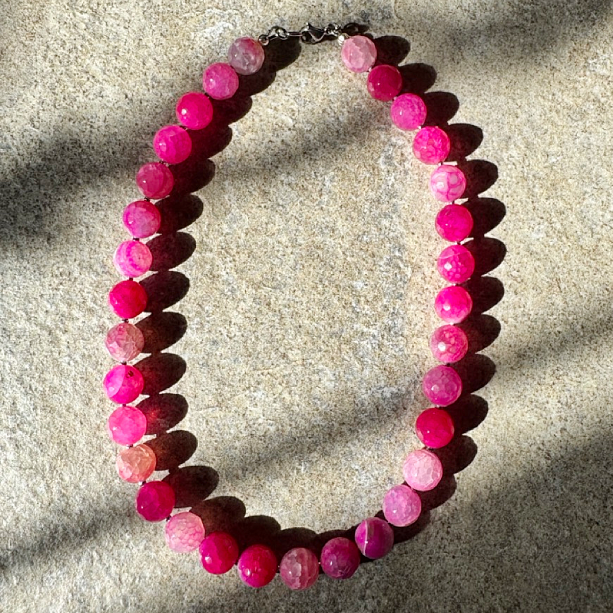 A beaded necklace made of round faceted pink gemstone beads in varying shades of magenta and rose, arranged in an oval on a textured beige stone surface with strong natural sunlight casting shadows.